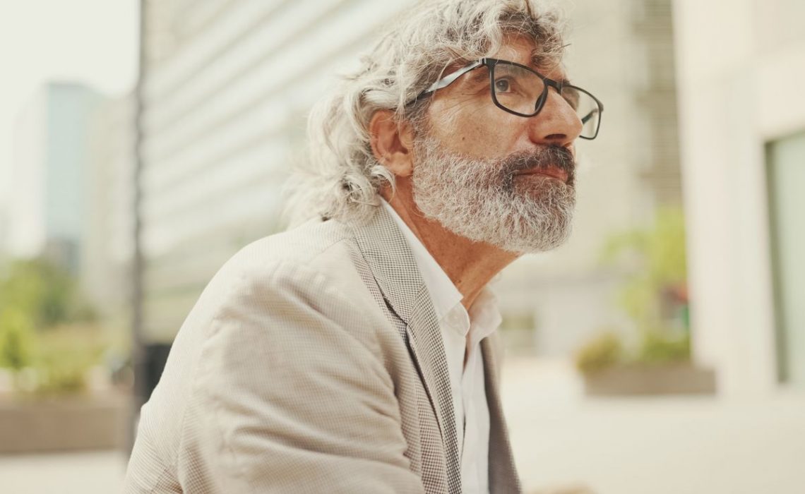 Clouse up, mature businessman with beard in eyeglasses wearing gray pondering jacket looks around