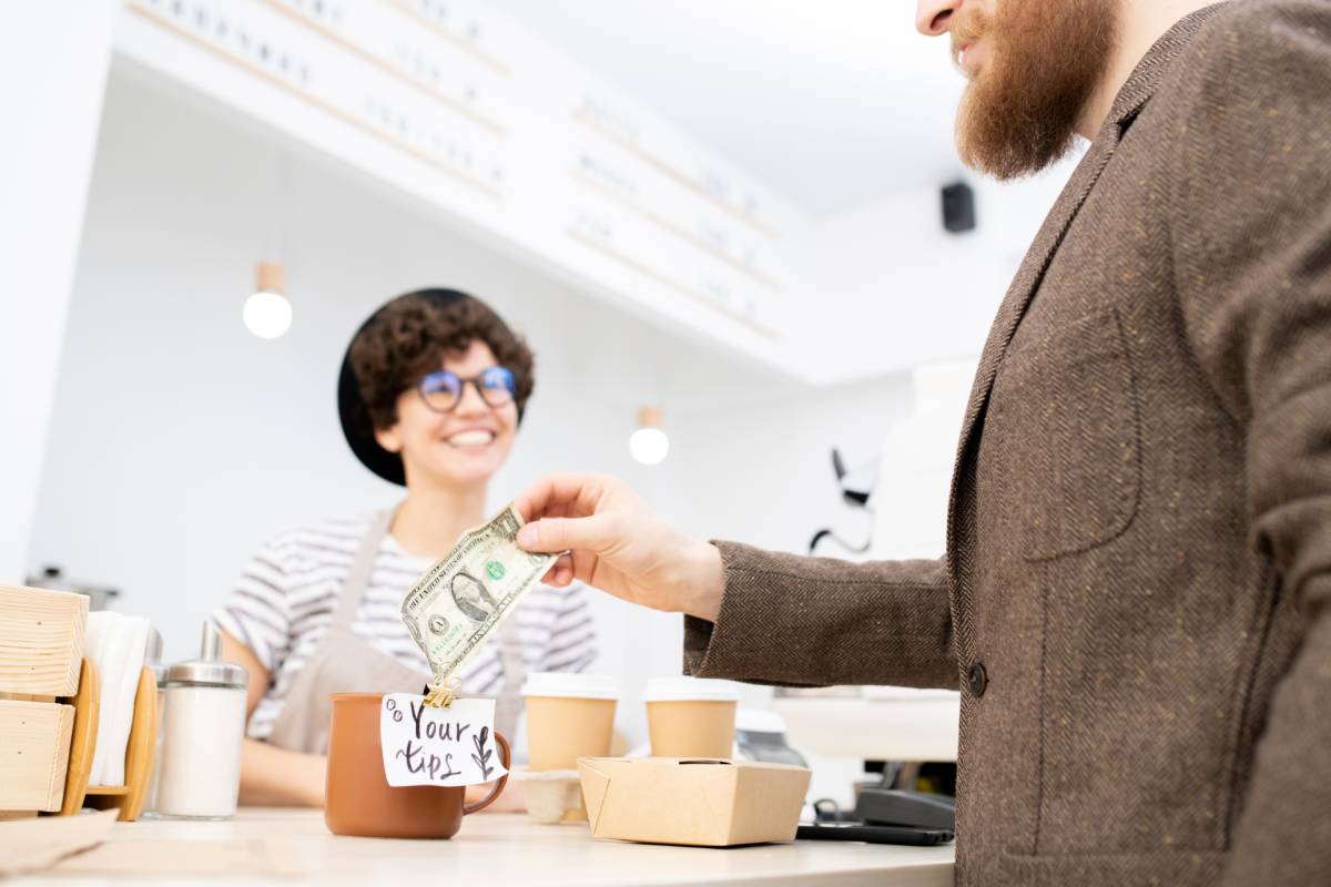 Close-up of bearded man in jacket leaving tip for friendly barista while buying coffee and snacks in coffee shop