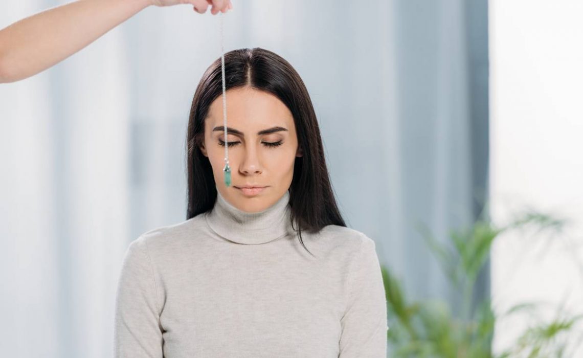 cropped shot of hypnotist with pendulum hypnotizing young woman with closed eyes