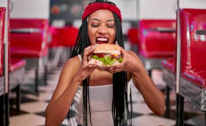 Young woman with braided hairstyle sitting on the floor and grabbing a big hamburger with her two ha