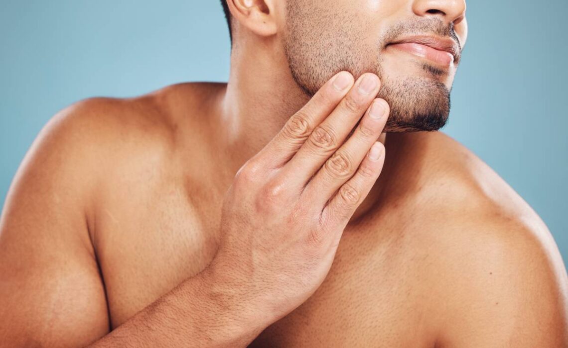 Man, beard care and hands on face after grooming, fresh and clean shave isolated on blue studio bac.
